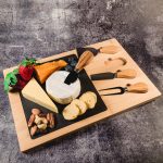 A bamboo cheese board featuring a slate surface, assorted cheeses, fruits, nuts, and cheese knives.
