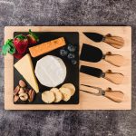 A bamboo cheese board featuring a variety of cheeses, fruits, nuts, and three wooden-handled knives.