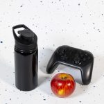 A drink bottle with a free flow lid, presented in sleek black, alongside a gaming controller and a red apple.
