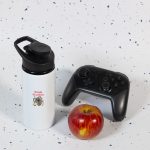 A drink bottle in white with a black lid, accompanied by a red apple and a black gaming controller.