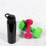 A drink bottle in black with a free flow lid, placed next to pink and green dumbbells on a speckled surface.