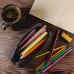 A gray felt utility case filled with colorful pens and pencils, alongside a notebook and a mug of coffee.