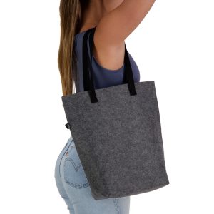 A grey tote bag with black handles, displayed by a person holding it by the straps. The bag has a logo.