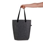 A hand holds a grey felt tote bag with black straps. The bag features a branding logo.