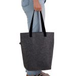 A grey tote bag with black handles, being held by a person wearing blue jeans. The bag has a logo.
