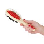 A pet grooming brush with a cream handle, red bristles, and black rubber base held in a hand with red nails.