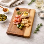 A wooden serving board featuring cured meats, cheese, crackers, olives, and fresh herbs, with a minimalist branding logo.