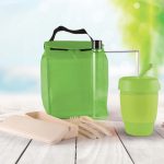 A green picnic bag contains a drink bottle, reusable cup, utensils, and food containers, set against a blurred outdoor background.