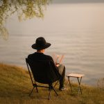 A black folding chair sits by the water, with a man in a hat reading a book beside a small table.