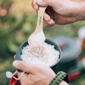 A hand holds a beige spoon scooping rice from a gray bowl outdoors. The spoon features a logo.