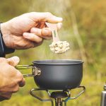 A hand holds a light-colored wooden utensil over a steaming gray pot, set against a natural background.