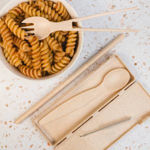 A tan Ramen Cutlery Set with a spoon, fork, and chopsticks, placed beside a bowl of rotini pasta.