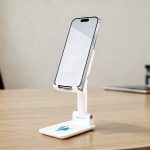 A white phone stand with an adjustable holder, displaying a smartphone on a wooden desk surface.