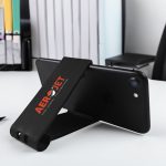 A black phone stand with a logo, positioned upright on a desk beside office supplies.