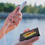 A black power bank with a logo is connected to a smartphone, held by a person near a body of water.