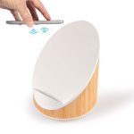 A wireless charger and speaker with a white top and bamboo base. A smartphone is held above the charger.