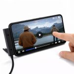 A smartphone displaying a video of a person by a lake, resting on a stand with a charging cable attached.