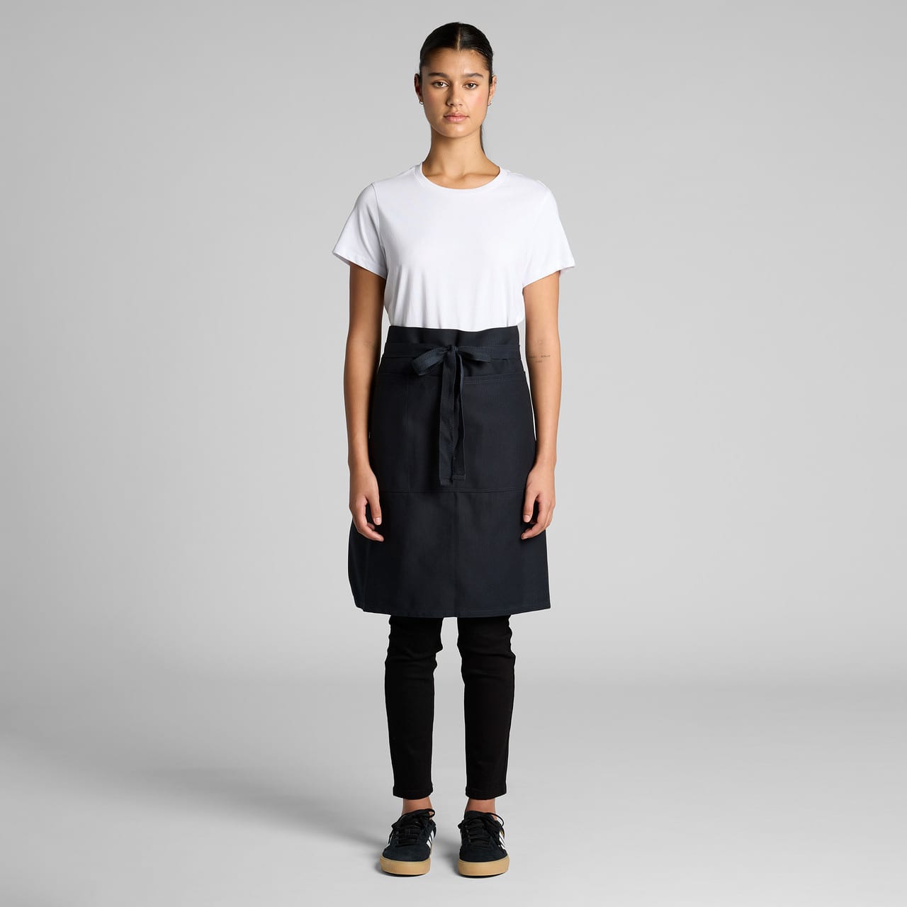 A half apron in black, featuring a front pocket and a tied waistband, displayed on a model wearing casual attire. A half apron in black, featuring a front pocket and a tied waistband, displayed on a model wearing casual attire.
