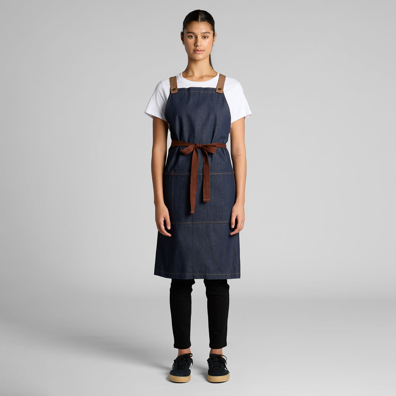 A denim apron featuring brown leather straps and a tie, displayed on a model against a neutral background. A denim apron featuring brown leather straps and a tie, displayed on a model against a neutral background.