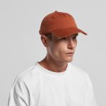 A five panel cap in a burnt orange colour, worn by a model, featuring a logo on the side.