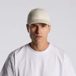 A beige cap worn by a model, featuring a front-panel design and a logo. The model is dressed in a white T-shirt.