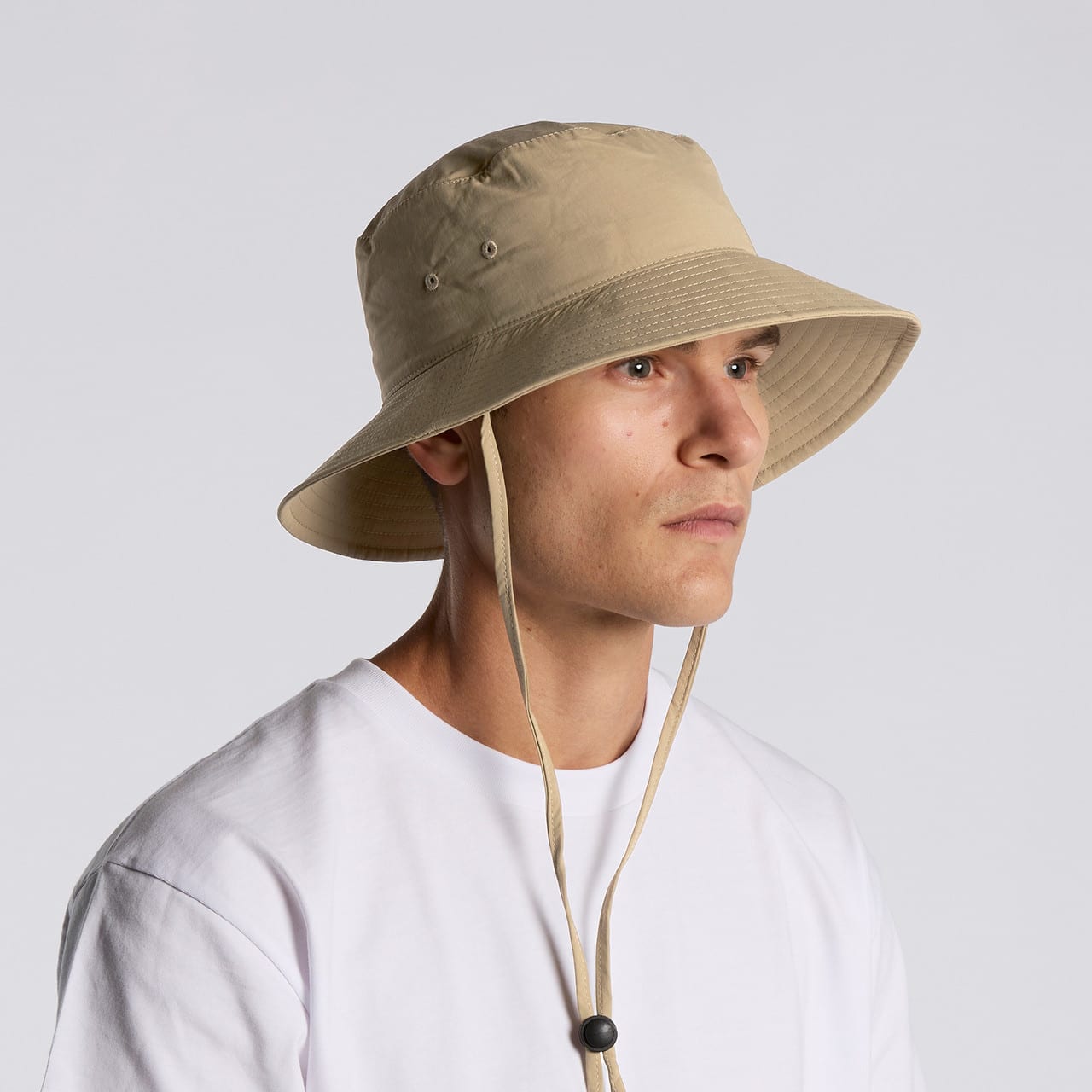 A nylon wide brim bucket hat in beige, featuring an adjustable chin strap and detailing around the brim. A nylon wide brim bucket hat in beige, featuring an adjustable chin strap and detailing around the brim.