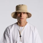A beige nylon wide brim bucket hat with a drawstring, featuring a logo, worn by a person in a white shirt.