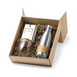A gift box containing a silver drink bottle and a glass with a wooden base, nestled in brown packing material.
