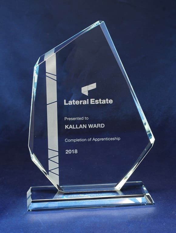 A glass award with a geometric design, featuring an engraved inscription and a base, resting on a blue background. Glass Sail - Image 1