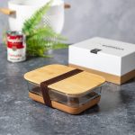 A glass lunch box with a bamboo lid and cork base, featuring a brown strap. Green foliage and a can are in the background.