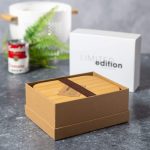 A bamboo lunch box in a kraft paper box with a brown strap, surrounded by greenery and a white container.