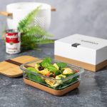 A glass lunch box with a bamboo lid contains pasta and fresh vegetables, set against a grey background.