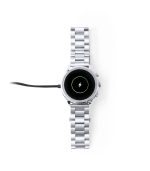 A silver smart watch with a circular face, charging with a cable attached, featuring a logo on the dial.