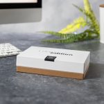 A white and brown box with a logo, placed on a grey surface surrounded by greenery and a computer keyboard.