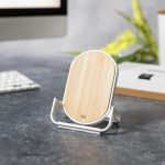A wireless charger with a wooden top and white base, positioned on a desk with a computer and greenery in the background.