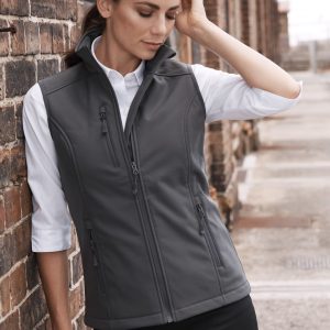 A woman in a fitted grey work vest over a white shirt stands against a brick wall. The vest has a logo on it.