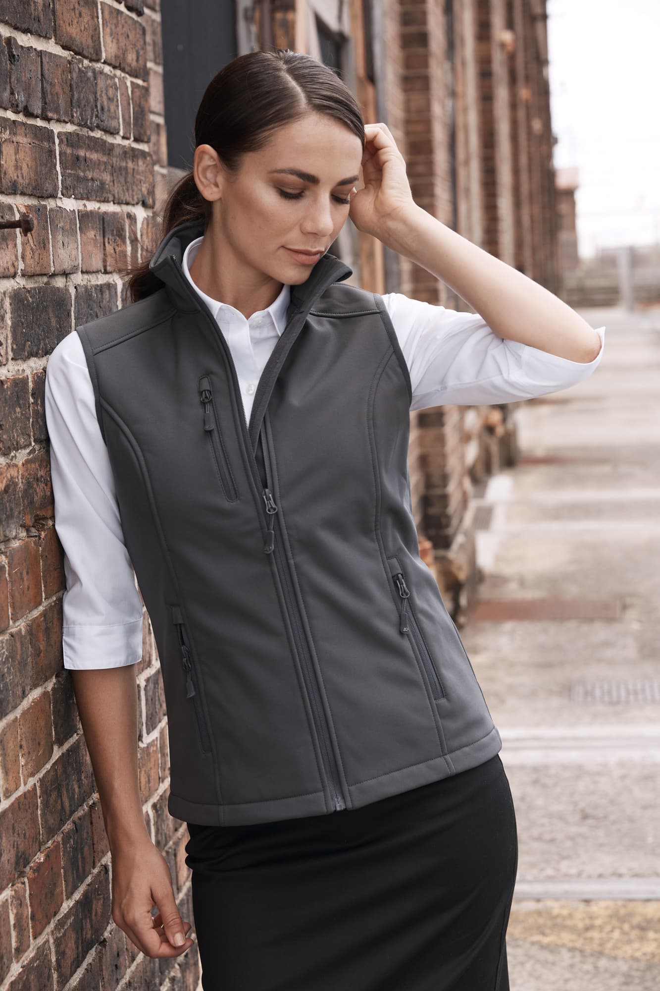 A woman in a fitted grey work vest over a white shirt stands against a brick wall. The vest has a logo on it. A woman in a fitted grey work vest over a white shirt stands against a brick wall. The vest has a logo on it.