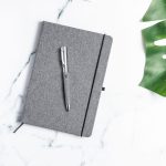 A grey notepad with a pen resting on top, surrounded by a marble surface and a green leaf.