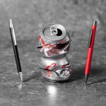 A red and a black metal pen stand beside a crushed silver soda can on a textured background.