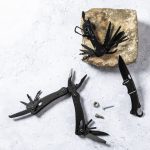 A black multitool with various tools extends from a stone, accompanied by a sleek black knife and screws.