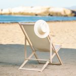 A leisure chair with a cream seat and wooden frame sits on the sand, topped with a wide-brimmed straw hat.