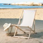 A beige leisure chair with a wooden frame beside a canvas bag and straw hat on a sandy beach with water in the background.