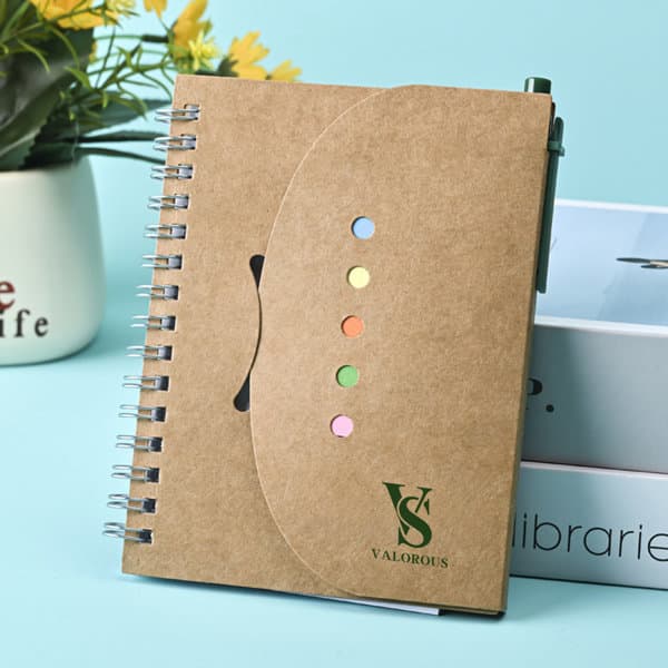 A brown spiral notebook with colourful sticky notes and a pen, featuring a logo on the cover. A brown spiral notebook with colourful sticky notes and a pen, featuring a logo on the cover.