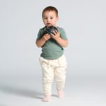 A toddler wears a green t-shirt and cream-coloured pants, holding a small ball in front of a light background.