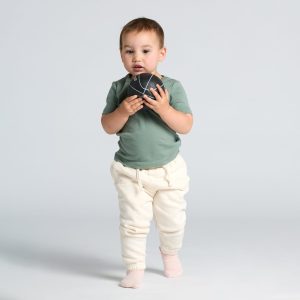 A toddler wears a green t-shirt and cream-coloured pants, holding a small ball in front of a light background.