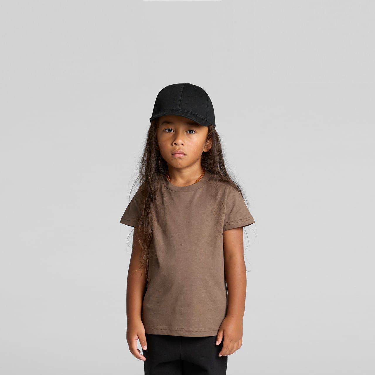 A brown kids' t-shirt displayed on a child wearing a black cap, set against a plain grey background. A brown kids' t-shirt displayed on a child wearing a black cap, set against a plain grey background.