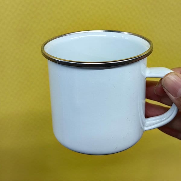 A white enamel campfire coffee mug with a gold rim, held by a hand, against a yellow background. A white enamel campfire coffee mug with a gold rim, held by a hand, against a yellow background.
