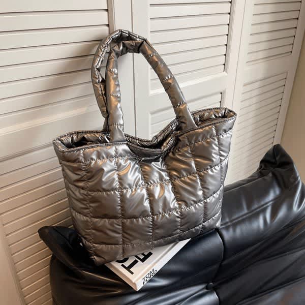 A large quilted tote bag in a shiny silver colour, featuring two handles, resting on a black sofa. A large quilted tote bag in a shiny silver colour, featuring two handles, resting on a black sofa.