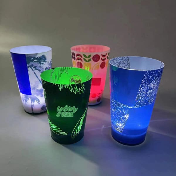 Four illuminated party cups in various designs and colours: green, blue, pink, and a patterned cup with a logo. Four illuminated party cups in various designs and colours: green, blue, pink, and a patterned cup with a logo.