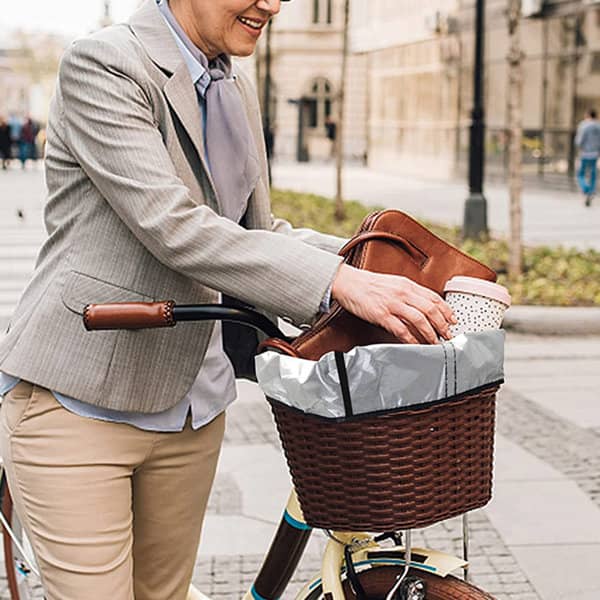 A waterproof bike basket liner in grey is being placed in a woven bike basket. A drink bottle and bag are visible. A waterproof bike basket liner in grey is being placed in a woven bike basket. A drink bottle and bag are visible.